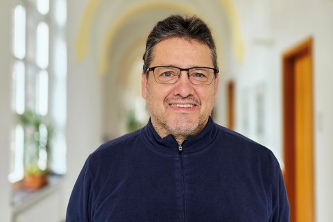 Ein Mann mit kurzem Haar und einer dunklen Brille steht lächelnd in einem Flur mit Gewölbedecke. Im Hintergrund sieht man ein  Fenster mit Blumenstock und eine Holztür.