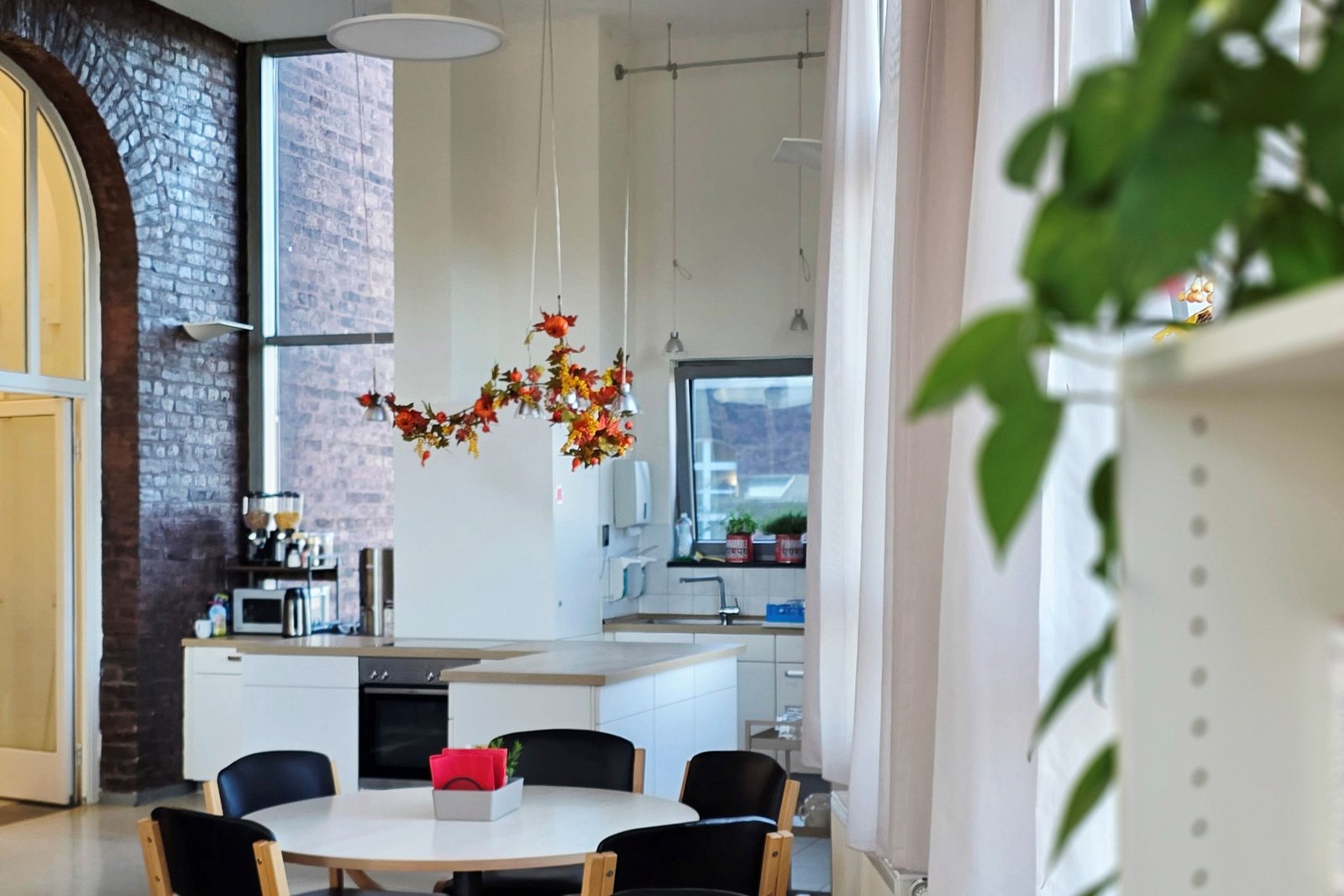 In einem hohen Raum mit Backstein-Sichtmauer sind Tische und Stühle umgeben von Pflanzen und einer Küchenzeile zu sehen. 