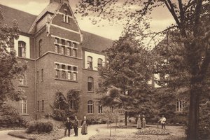 Ein sepiafarbenes historisches Foto aus den Anfängen des neugebauten Klosters der Alexianer Brüder in Köln Ensen. Ein großer Backsteinbau mit großen Bäumen und drei Personen im Vordergrund.