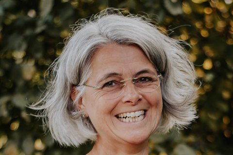 Eine Frau mit grauen Haaren und Bobfrisur steht lächelnd vor einem grünen Busch im Freien.