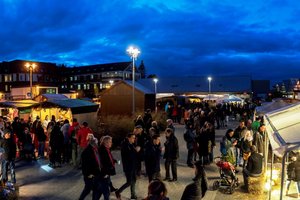 Unser Alexianer Weihnachtsmarkt ist in jedem Jahr ein Besuchermagnet.  Unter nachtblauem Himmel ist ein Platz mit vielen erleuchteten Holzbuden und im Vordergrund ein erleuchtetes Zelt mit Stroh auf dem Boden zu sehen, an denen viele Besucher entlang gehen.