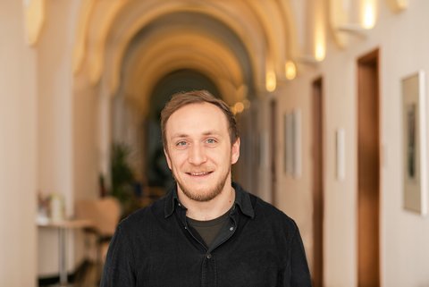 Ein Mann mit schwarzem Hemd und schwarzem TShirt steht lächelnd in einem erleuchteten Flur mit Gewölbedecke.