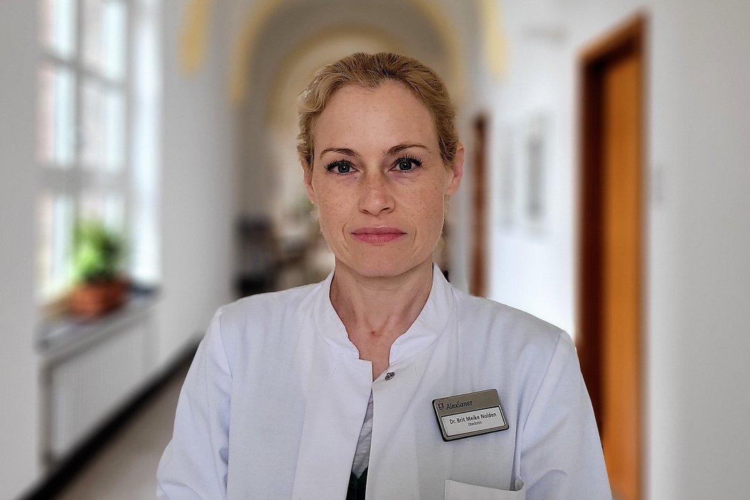 Dr. Brit Nolden Eine blonde Frau schaut in einem weißen Arztkittel lächelnd in die Kamera auf einem Flur mit Gewölbedecke und großen Fenstern.