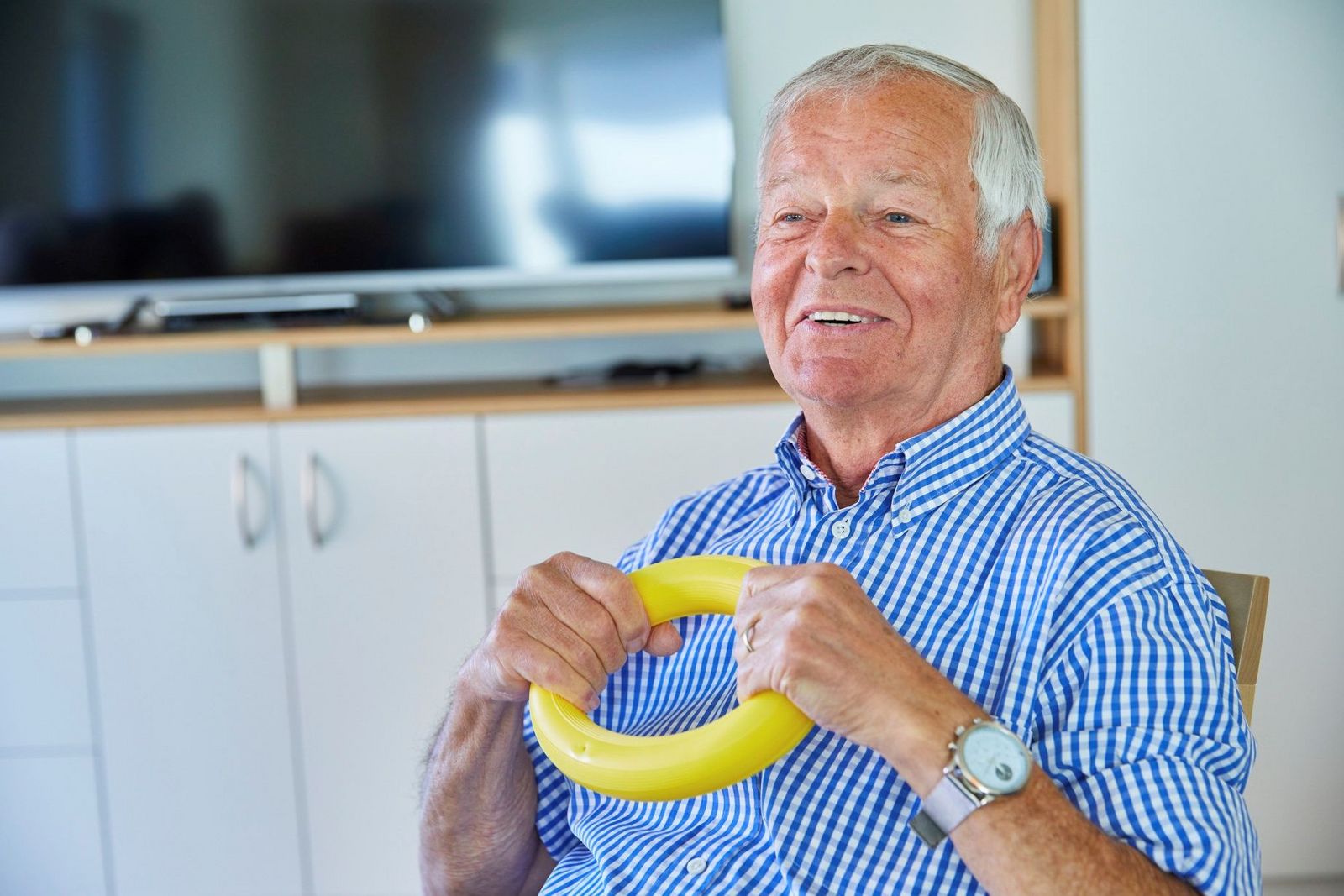 Ein älterer Mann in einem blauweißgestreiften Hemd mit kurzen Ärmeln hält einen gelben Plastikring mit beiden Händen und lacht dabei.