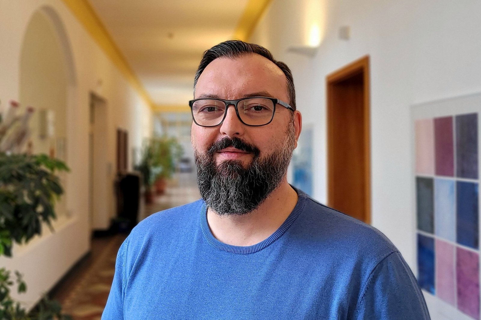 Mann mit Brille und Vollbart steht in einem beleuchteten Krankenhausflur mit Bildern und Fenstern.
