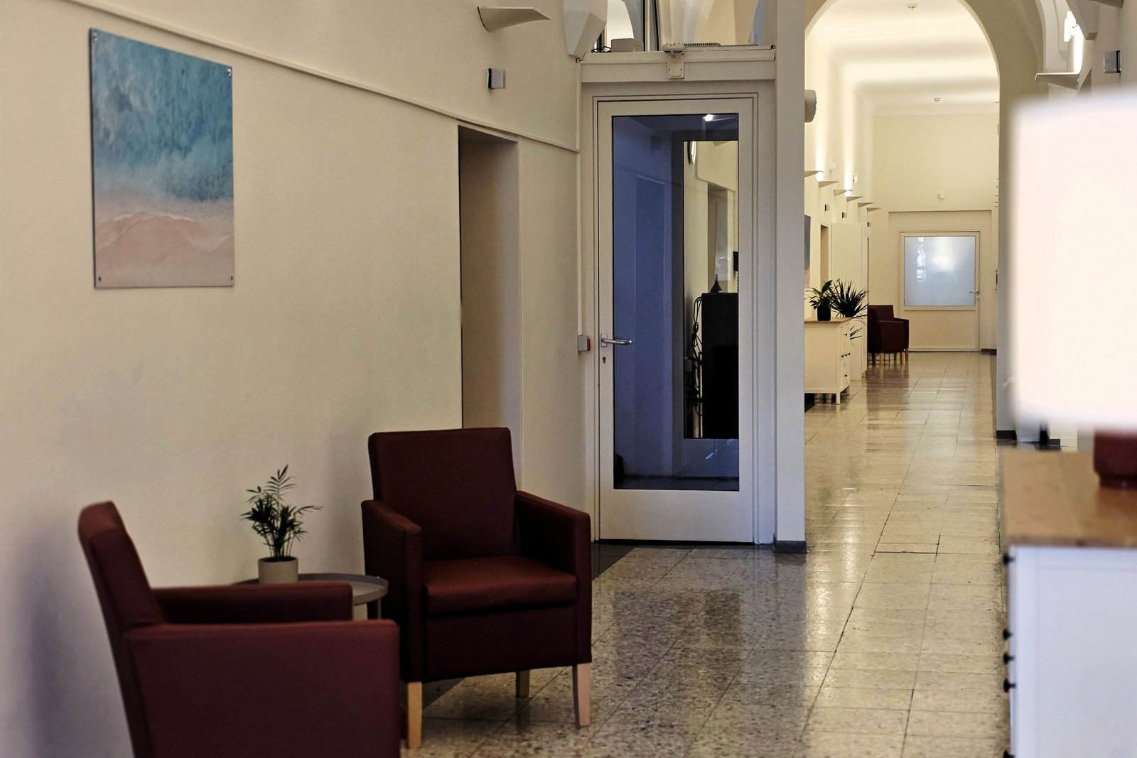 Elegante Sitzledermöbel stehen an der Wand undter einem Kunstwerk. Eine Stehlampe ist erleuchtet und der Blick richtet sich auf einen Flur mit einer nach oben rundbogengeformten Öffnung. 