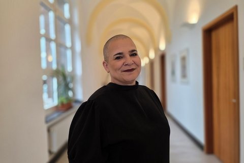 Ein Frau mit ganz kurzen Haaren und einem schwarzen Pulli steht lächelnd in einem hellen Flur mit Gewölbedecke und Fenstern und Holztüren im Hintergrund.